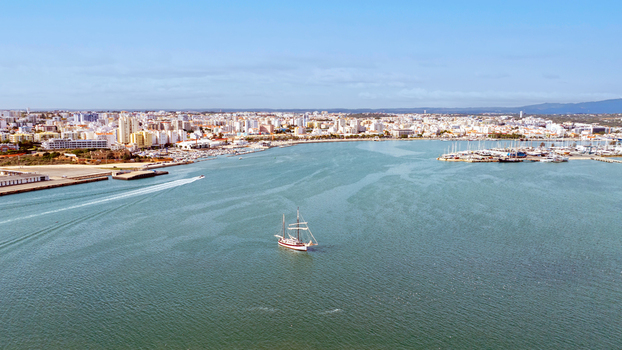 Aerial view of a sailboat sailing in the harbor with the coastal cityscape and marina in the background in Portimao Faro District Portugal. Aerial view of a sailboat sailing in the harbor with the coastal cityscape and marina in the background in Portimao Faro District Portugal.