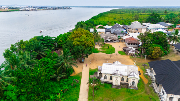 Aerial view of a church surrounded by lush trees and residential buildings near a wide river with boats in Minima Rivers Nigeria.