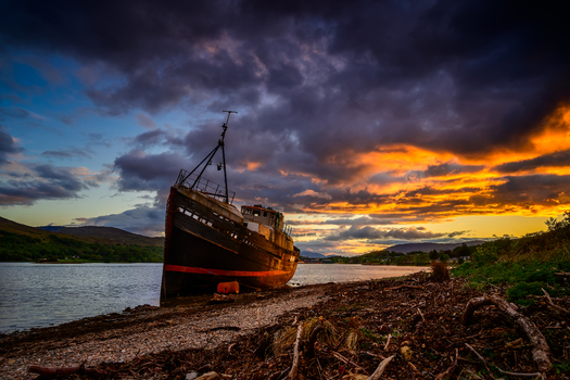 View of an old shipwreck resting on the rocky shore under a dramatic sunset sky with dark clouds and golden light in Isle of Skye Scotland. View of an old shipwreck resting on the rocky shore under a dramatic sunset sky with dark clouds and golden light in Isle of Skye Scotland.