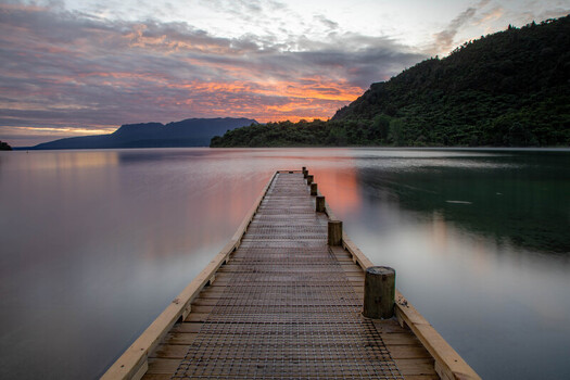 View of Lake Tarawera with a wooden jetty extending into the calm water under a vibrant sunset sky in Rotorua Bay of Plenty Region New Zealand. View of Lake Tarawera with a wooden jetty extending into the calm water under a vibrant sunset sky in Rotorua Bay of Plenty Region New Zealand.