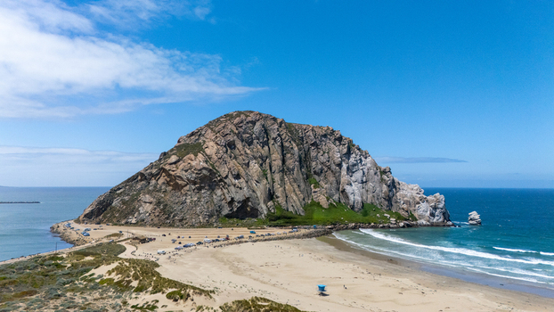 Aerial view of Morro Rock standing prominently over the sandy beach and blue ocean waters under a clear sky in Morro Bay California United States.