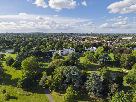 Aerial view of Gunnersbury Park featuring the historic mansion surrounded by lush green lawns mature trees and a small lake under a blue sky in London England United Kingdom. Aerial view of Gunnersbury Park featuring the historic mansion surrounded by lush green lawns mature trees and a small lake under a blue sky in London England United Kingdom.