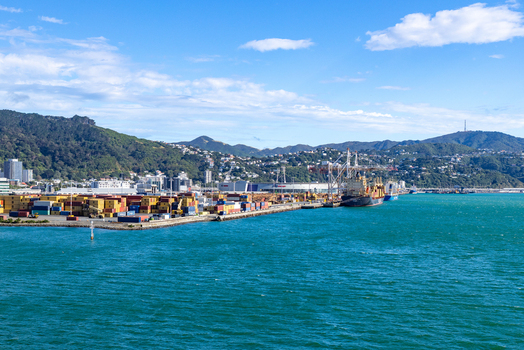 Wellington New Zealand 18 February 2026 View of containers stacked high on the wharf ships docked along the pier set against the backdrop of verdant hills and a bright sky.