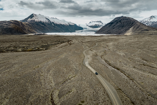 Aerial view of a car driving on a winding dirt road through a vast barren volcanic landscape with the Vatnajokull glacier and snow capped mountains in the background Iceland.