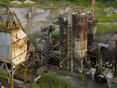 Aerial view of an abandoned industrial site featuring rusted metal silos processing machinery and weathered structures set within a grassy landscape in Poland. Aerial view of an abandoned industrial site featuring rusted metal silos processing machinery and weathered structures set within a grassy landscape in Poland.