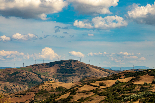 View of wind turbines on a rolling hill ridge surrounded by dry Mediterranean landscape under a blue sky in Sardinia Italy.