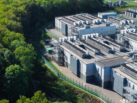 Aerial view of a modern data center facility featuring rows of industrial cooling units on the roof enclosed by a security fence and bordered by a dense forest in the United Kingdom.