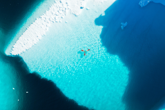 Aerial view of three people in red swimsuits swimming in crystal clear turquoise water beside a massive white iceberg in Greenland. Aerial view of three people in red swimsuits swimming in crystal clear turquoise water beside a massive white iceberg in Greenland.
