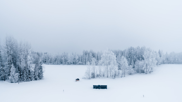 Aerial view of a snow covered landscape with a frost covered forest and a small cabin in the middle of a vast white field in Latvia. Aerial view of a snow covered landscape with a frost covered forest and a small cabin in the middle of a vast white field in Latvia.