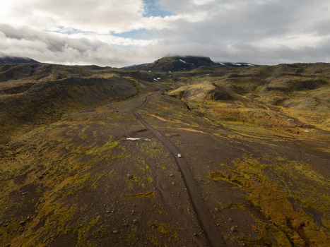 Aerial view of a solitary white car navigates the rugged moss covered terrain beneath a cloudy sky leading towards distant snow capped mountains Snæfellsnes Iceland. Aerial view of a solitary white car navigates the rugged moss covered terrain beneath a cloudy sky leading towards distant snow capped mountains Snæfellsnes Iceland.