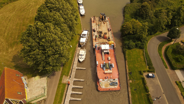 Aerial view of a large work barge navigating a canal alongside smaller moored boats with lush green banks trees and a nearby road in Saint Omer Hauts de France France. Aerial view of a large work barge navigating a canal alongside smaller moored boats with lush green banks trees and a nearby road in Saint Omer Hauts de France France.