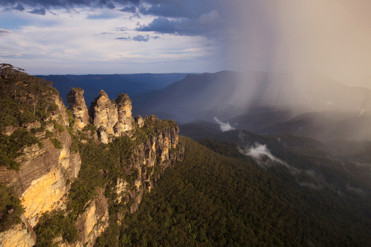 View of the Three Sisters rock formation overlooking a deep forested valley with a rain shaft under a dramatic sky in Australia. View of the Three Sisters rock formation overlooking a deep forested valley with a rain shaft under a dramatic sky in Australia.