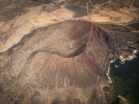Aerial view of a volcanic crater with rugged red and black rock formations surrounded by arid terrain and the coastline of Lanzarote Canarias Spain. Aerial view of a volcanic crater with rugged red and black rock formations surrounded by arid terrain and the coastline of Lanzarote Canarias Spain.