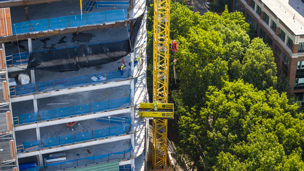 Aerial view of Kendall Square construction site with a yellow tower crane and workers on building levels surrounded by lush green trees in Cambridge Massachusetts United States. Aerial view of Kendall Square construction site with a yellow tower crane and workers on building levels surrounded by lush green trees in Cambridge Massachusetts United States.