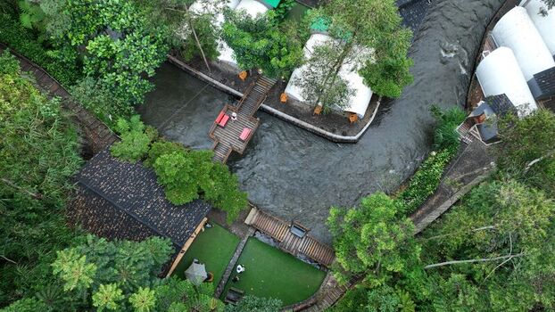 Aerial view of a glamping resort featuring white tent structures wooden footbridges over a flowing river lush green trees and manicured lawns in Pangalengan West Java Indonesia. Aerial view of a glamping resort featuring white tent structures wooden footbridges over a flowing river lush green trees and manicured lawns in Pangalengan West Java Indonesia.