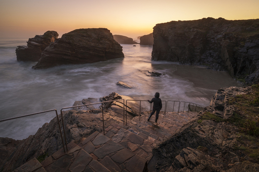 View of Praia das Catedrais with a person standing on stone stairs overlooking dramatic rock formations and sea arches during a golden sunset in Lugo Lugo Spain. View of Praia das Catedrais with a person standing on stone stairs overlooking dramatic rock formations and sea arches during a golden sunset in Lugo Lugo Spain.