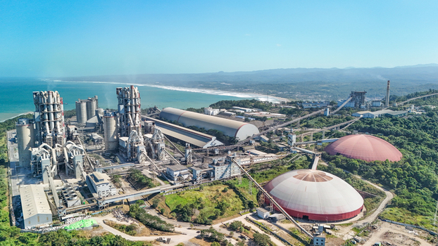 Aerial view of a large industrial cement plant with tall silos conveyor systems and dome shaped storage structures situated along a scenic coastline in Indonesia.