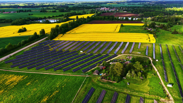 Aerial view of a solar farm with rows of photovoltaic panels situated amidst vibrant yellow rapeseed fields and green meadows in the rural countryside of Latvia. Aerial view of a solar farm with rows of photovoltaic panels situated amidst vibrant yellow rapeseed fields and green meadows in the rural countryside of Latvia.
