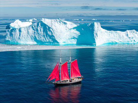 Aerial view of a sailboat with vibrant red sails navigating the deep blue waters near a massive towering iceberg in the Arctic Greenland. Aerial view of a sailboat with vibrant red sails navigating the deep blue waters near a massive towering iceberg in the Arctic Greenland.