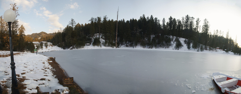 View of frozen Banjosa Lake surrounded by snow covered pine trees and mountains under a clear sky with a small boat on the shore in Rawalakot Kashmir Pakistan.