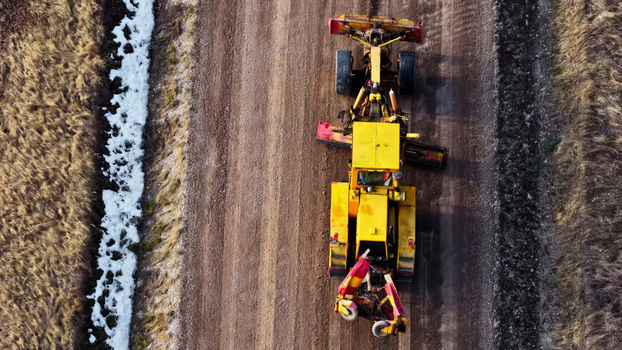 Aerial view of a yellow motor grader leveling a dirt road with patches of snow on the roadside Latvia. Aerial view of a yellow motor grader leveling a dirt road with patches of snow on the roadside Latvia.