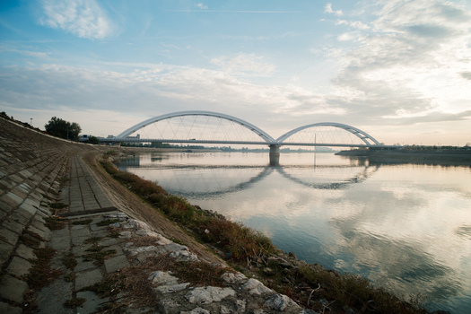 View of Zezelj Bridge spanning the Danube River with a stone embankment in the foreground under a soft sunset sky in Novi Sad Vojvodina Serbia. View of Zezelj Bridge spanning the Danube River with a stone embankment in the foreground under a soft sunset sky in Novi Sad Vojvodina Serbia.