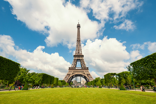 View of the Eiffel Tower rising above manicured green lawns and trimmed trees under a bright blue sky with fluffy white clouds in Paris Ile de France France.