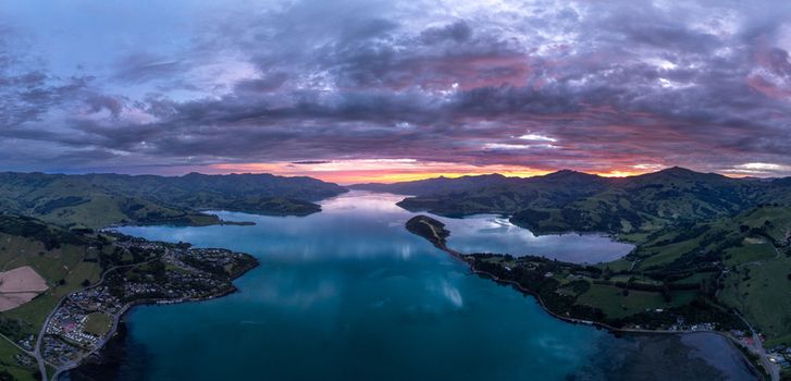 Aerial view of the coastal landscape of Akaroa Harbour with rolling green hills calm turquoise waters and a dramatic sunset sky in Duvauchelle Canterbury Region New Zealand. Aerial view of the coastal landscape of Akaroa Harbour with rolling green hills calm turquoise waters and a dramatic sunset sky in Duvauchelle Canterbury Region New Zealand.