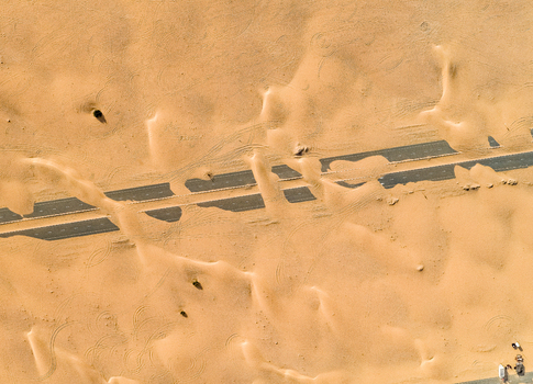 View of a paved road being overtaken by shifting orange sand dunes with visible tire tracks in the desert in Dubai Dubai United Arab Emirates. View of a paved road being overtaken by shifting orange sand dunes with visible tire tracks in the desert in Dubai Dubai United Arab Emirates.