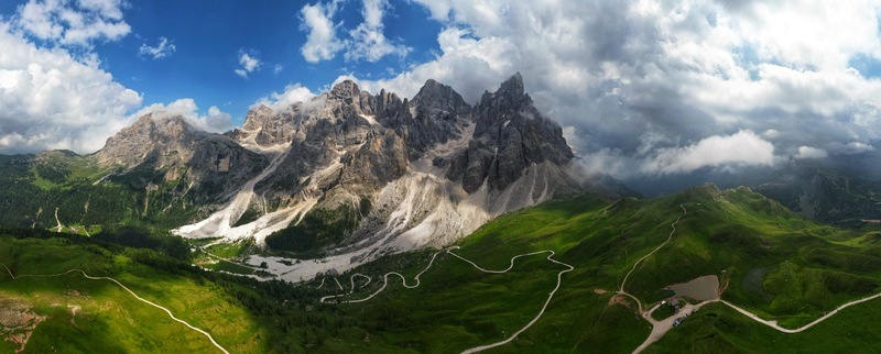 Aerial view of the Dolomite Mountains with jagged peaks lush green alpine meadows and winding roads under a dramatic cloudy sky in Canazei Trentino South Tyrol Italy. Aerial view of the Dolomite Mountains with jagged peaks lush green alpine meadows and winding roads under a dramatic cloudy sky in Canazei Trentino South Tyrol Italy.