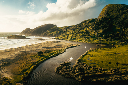 Aerial view of a kite surfer gliding along the river at Kapowairua Spirits Bay Campsite with rolling green hills and sandy coastline in Cape Reinga Northland Region New Zealand. Aerial view of a kite surfer gliding along the river at Kapowairua Spirits Bay Campsite with rolling green hills and sandy coastline in Cape Reinga Northland Region New Zealand.