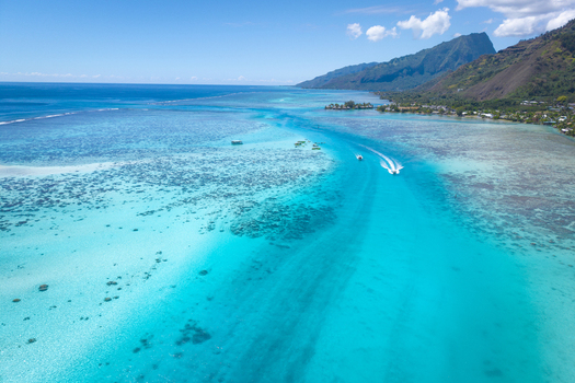 View of the turquoise lagoon with a boat leaving a wake surrounded by coral reefs and lush mountains in Moorea Maiao Windward Islands French Polynesia.