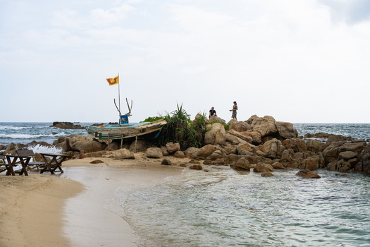 View of Secret Beach Mirissa featuring a traditional boat with a Sri Lankan flag on a rocky outcrop and people standing by the ocean in Mirissa Southern Province Sri Lanka. View of Secret Beach Mirissa featuring a traditional boat with a Sri Lankan flag on a rocky outcrop and people standing by the ocean in Mirissa Southern Province Sri Lanka.