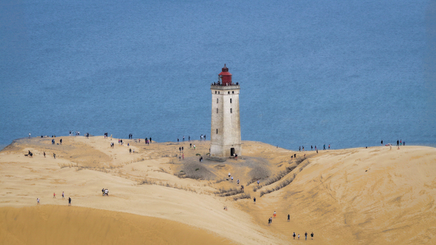 Aerial view of Rubjerg Knude lighthouse standing on sandy dunes with people walking nearby and the blue sea in the background Hjorring Municipality Denmark. Aerial view of Rubjerg Knude lighthouse standing on sandy dunes with people walking nearby and the blue sea in the background Hjorring Municipality Denmark.