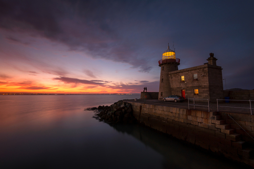 View of Howth Harbour lighthouse at sunset with a dramatic sky over calm waters and people standing on the pier in Ireland. View of Howth Harbour lighthouse at sunset with a dramatic sky over calm waters and people standing on the pier in Ireland.