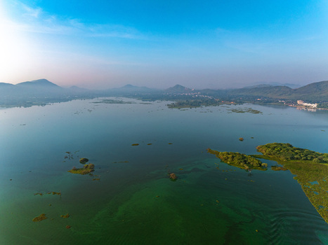 Aerial view of a serene lake with lush green islands and distant hills under a clear blue sky in Udaipur Rajasthan India. Aerial view of a serene lake with lush green islands and distant hills under a clear blue sky in Udaipur Rajasthan India.