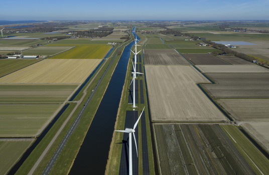 Aerial view of a row of modern wind turbines and solar panels along a straight canal through flat agricultural fields under a clear blue sky in Netherlands. Aerial view of a row of modern wind turbines and solar panels along a straight canal through flat agricultural fields under a clear blue sky in Netherlands.
