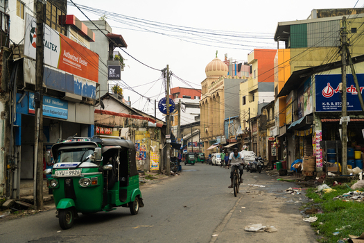 Sri Lanka 15 February 2026 View of a green tuk tuk and a cyclist on a narrow urban street lined with commercial shops and a mosque dome under an overcast sky.