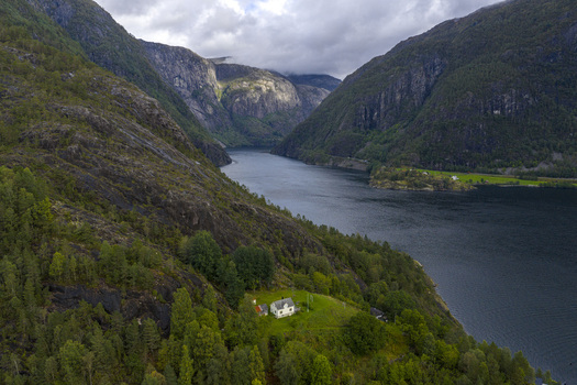 Aerial view of a small white house on a green hillside overlooking a deep fjord surrounded by steep rocky mountains under a cloudy sky Fjaera Vestland Norway. Aerial view of a small white house on a green hillside overlooking a deep fjord surrounded by steep rocky mountains under a cloudy sky Fjaera Vestland Norway.