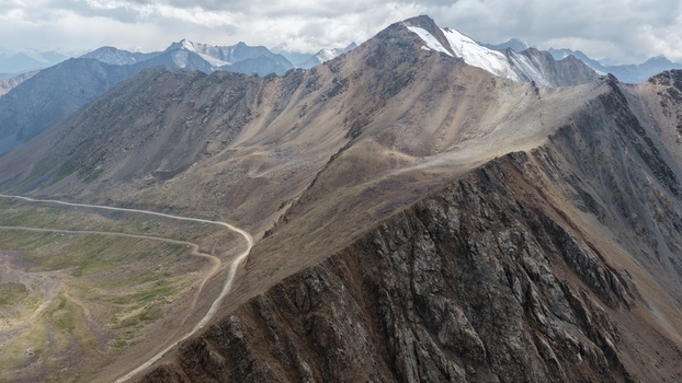 Aerial view of the rugged Enilchek mountain range in the Tian Shan featuring a winding dirt road cutting through rocky terrain under a cloudy sky in Enilchek Issyk Kul Region Kyrgyzstan. Aerial view of the rugged Enilchek mountain range in the Tian Shan featuring a winding dirt road cutting through rocky terrain under a cloudy sky in Enilchek Issyk Kul Region Kyrgyzstan.