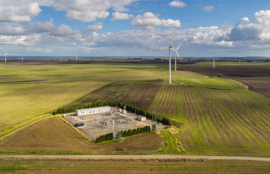 Aerial view of a wind farm and an electrical substation situated amidst vast agricultural fields under a cloudy blue sky in Pomeranian Voivodeship Poland.