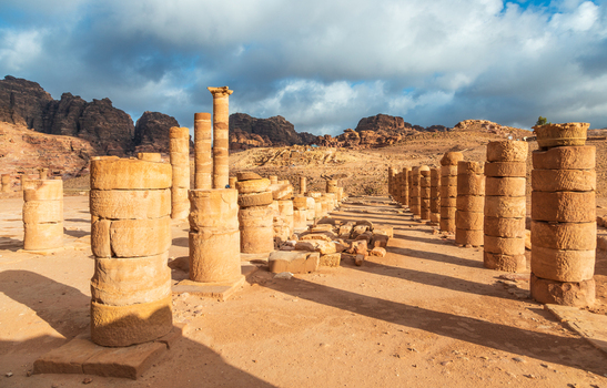View of the ancient Colonnaded Street with weathered stone columns and rocky desert mountains under a dramatic cloudy sky in Wadi Musa Ma an Governorate Jordan. View of the ancient Colonnaded Street with weathered stone columns and rocky desert mountains under a dramatic cloudy sky in Wadi Musa Ma an Governorate Jordan.