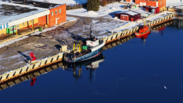 Aerial view of fishing boats docked at a snowy harbor pier next to red brick industrial buildings in Latvia. Aerial view of fishing boats docked at a snowy harbor pier next to red brick industrial buildings in Latvia.