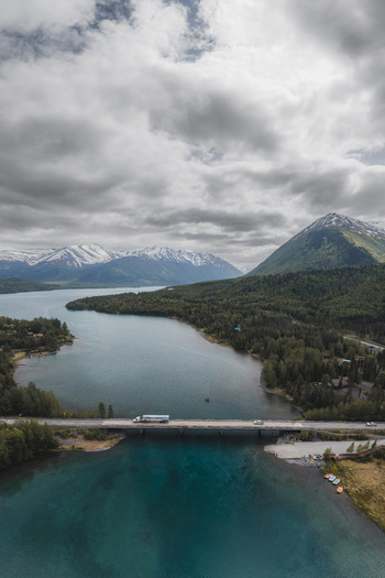 Aerial view of Alaska Wilderness featuring a bridge crossing a turquoise lake surrounded by dense pine forests and snow capped mountains under a cloudy sky in Palmer Alaska United States. Aerial view of Alaska Wilderness featuring a bridge crossing a turquoise lake surrounded by dense pine forests and snow capped mountains under a cloudy sky in Palmer Alaska United States.