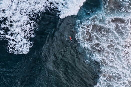 Aerial view of a surfer paddling through dark blue waves and white sea foam at Praia do Areal de Santa Barbara Ribeira Grande Acores Portugal. Aerial view of a surfer paddling through dark blue waves and white sea foam at Praia do Areal de Santa Barbara Ribeira Grande Acores Portugal.