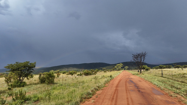 Aerial view of a red dirt road cutting through the green savanna landscape under a dramatic stormy sky with rolling hills and a faint rainbow Waterberg District Municipality Limpopo South Africa. Aerial view of a red dirt road cutting through the green savanna landscape under a dramatic stormy sky with rolling hills and a faint rainbow Waterberg District Municipality Limpopo South Africa.
