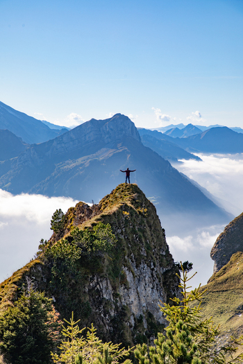 Aerial view of a person standing with arms outstretched on a rocky mountain peak in the Swiss Alps surrounded by clouds and mountain ranges in Airolo Ticino Switzerland. Aerial view of a person standing with arms outstretched on a rocky mountain peak in the Swiss Alps surrounded by clouds and mountain ranges in Airolo Ticino Switzerland.