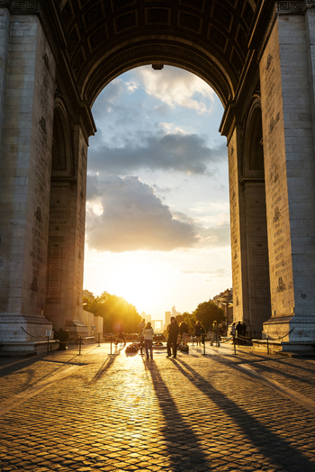 View of the Arc de Triomphe at sunset with people walking on the cobblestone pavement and long shadows stretching across the ground in Paris Ile de France France.