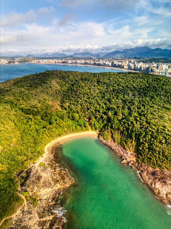 Aerial view of Praia do Ermitao featuring a secluded beach with turquoise water surrounded by lush green hills and rocky cliffs with a distant skyline Guarapari Espirito Santo Brazil. Aerial view of Praia do Ermitao featuring a secluded beach with turquoise water surrounded by lush green hills and rocky cliffs with a distant skyline Guarapari Espirito Santo Brazil.