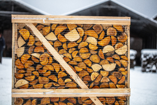 View of a wooden crate filled with stacked firewood wrapped in plastic netting in a snowy outdoor setting in Latvia. View of a wooden crate filled with stacked firewood wrapped in plastic netting in a snowy outdoor setting in Latvia.
